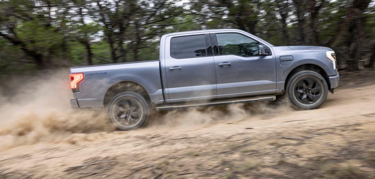 The side profile of a Ford F-150 Lightning Lartiat electric full-size pickup truck driving on a dusty road