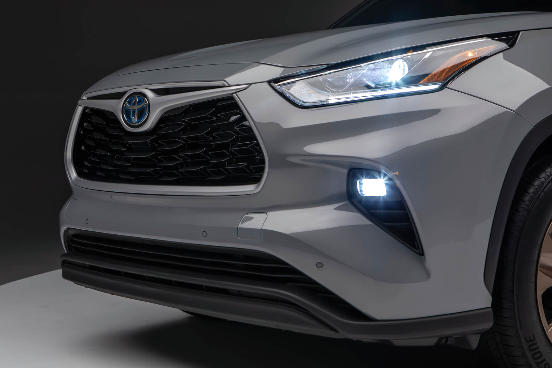 Front diagonal view of a grey 2023 Highlander Hybrid with headlamps and fog lamps illuminated. This model is one of the best Toyota hybrid models