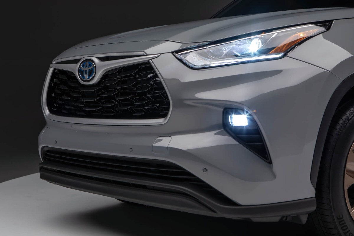 Front diagonal view of a grey 2023 Highlander Hybrid with headlamps and fog lamps illuminated.