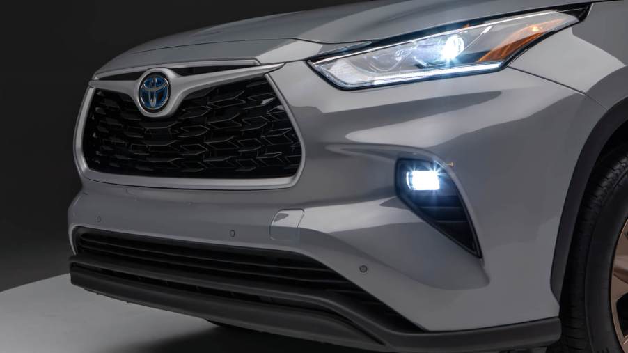 Front diagonal view of a grey 2023 Highlander Hybrid with headlamps and fog lamps illuminated. This model is one of the best Toyota hybrid models