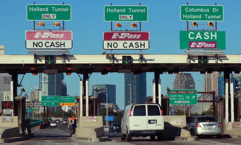 A van and silver sedan drive through a toll plaza to the Hollan tunnel, the skyline of New York City visible in the background.