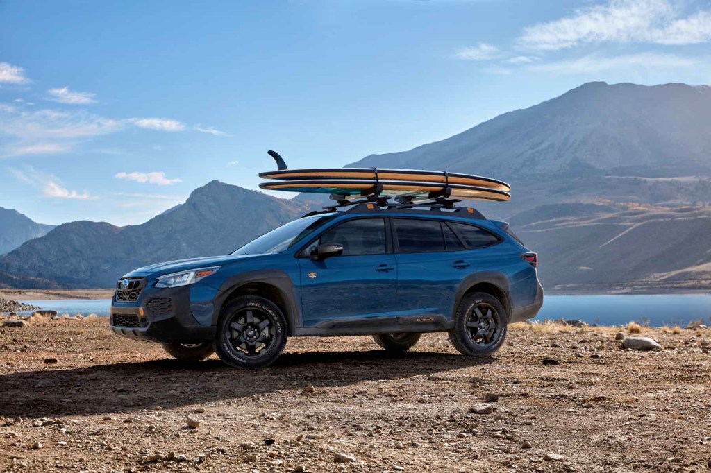 Wether you call it an SUV or Wagon, this blue Subaru Outback wilderness sits in a desert and mountain landscape with a surfboard on its roof racks. 