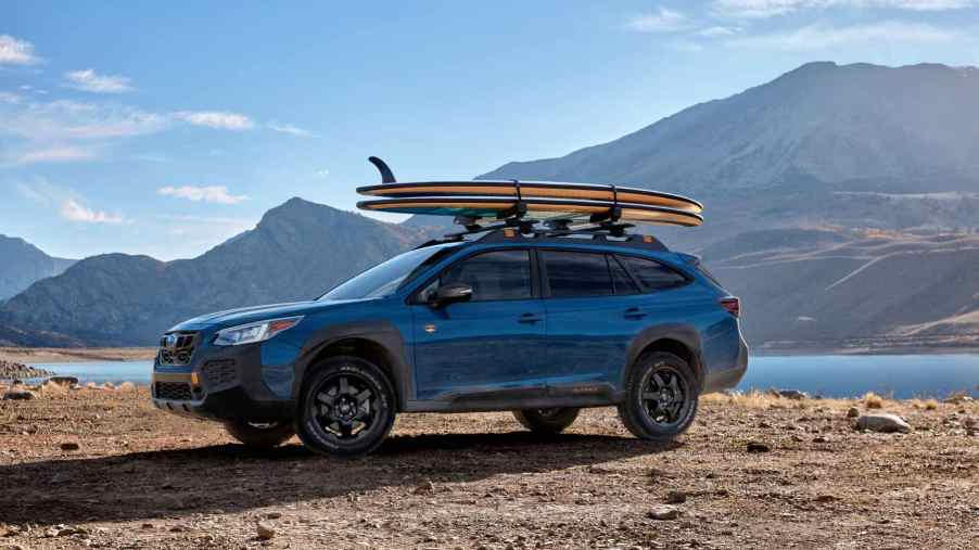 Wether you call it an SUV or Wagon, this blue Subaru Outback wilderness sits in a desert and mountain landscape with a surfboard on its roof racks.