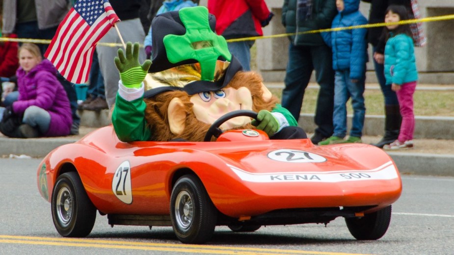 Leprechaun driving a tiny orange car in a St. Patrick’s Day parade 
