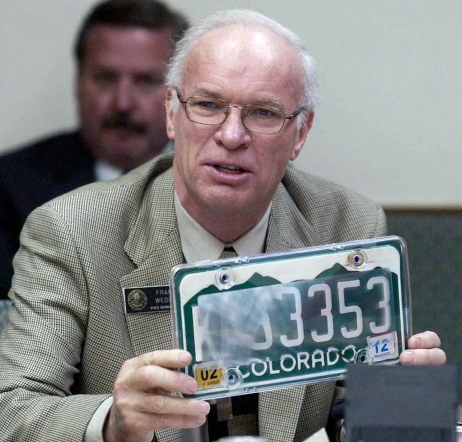 A man in a suit holds up a license plate with a clear tape cover designed to trick automatic readers.