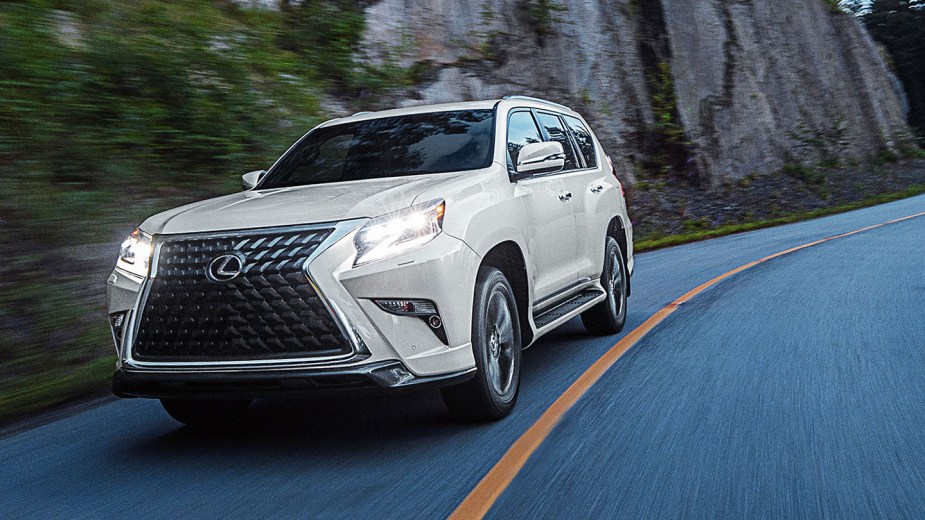 A white 2023 Lexus GX SUV drives toward the camera with its headlamps on.