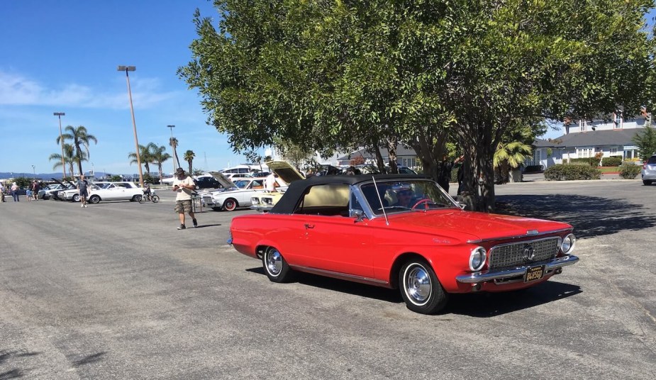 A bright red Plymouth Valiant classic car rolls though a car show, a row of slant-six Dodge Darts and other cars visible in the background.