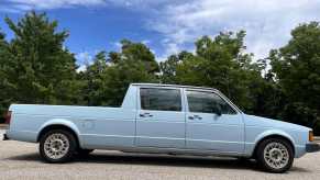 A blue 1981 Volkswagen Rabbit pickup that someone stretched into a sort of midsize VW truck, parked on pavement with trees in the background.
