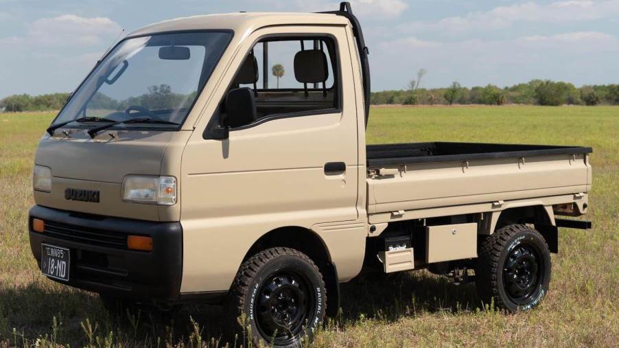 A tan Suzuki Carry "Kei-Class" mini truck parked in a field, trees visible in the background.