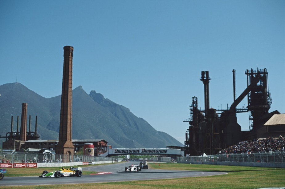 Grand Prix cars circle a race track in front of factory smokestacks in Monterrey Mexico where Tesla is building a plant.