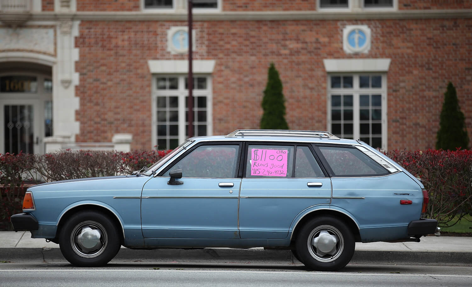 A blue station wagon parked on the street with an $1100 sign and phone number for used car buyers, a brick house in the background.