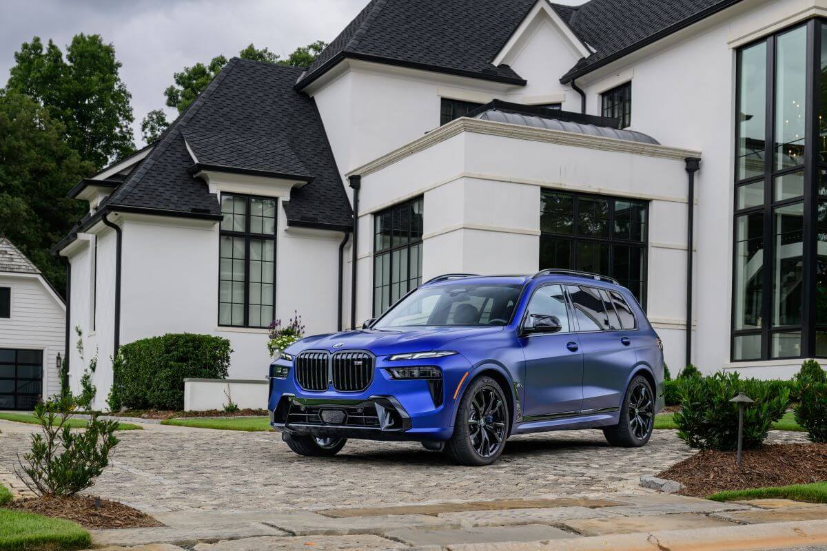 A blue 2023 BMW X7 full-size luxury SUV model parked on a cobblestone driveway outside a home
