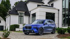 A blue 2023 BMW X7 full-size luxury SUV model parked on a cobblestone driveway outside a home