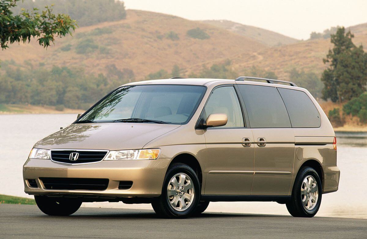 A gold/beige 2003 Honda Odyssey minivan model parked at the edge of a forest lake