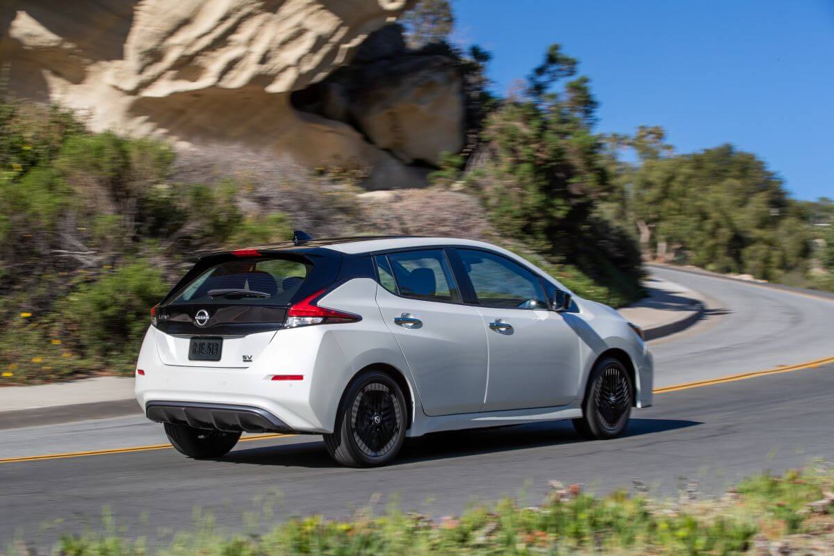 A rear side profile shot of a white 2023 Nissan Leaf all-electric hatchback driving up a curving hill road