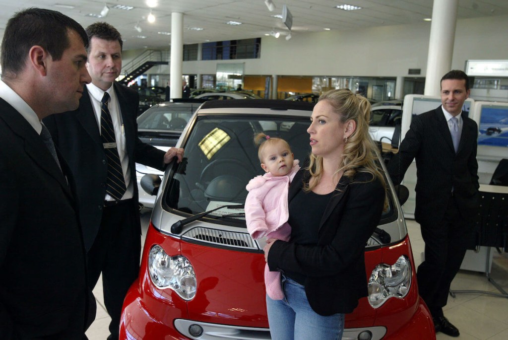A woman and her baby are surrounded by salesmen when shopping for a car.