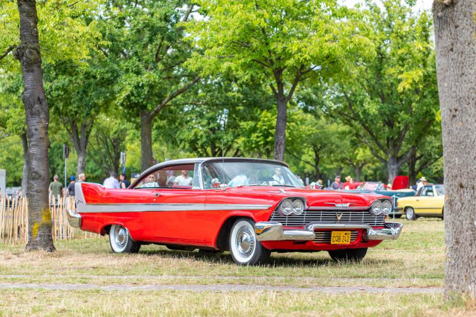A red 1958 Plymouth Fury from the movie Christine is parked at a car show, trees visible in the background.