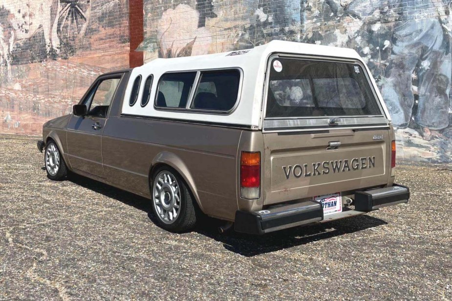 The rear of a VW Rabbit pickup truck with its white Leer cap and the Volkswagen lettering on its tailgate visible, parked in front of a brick wall painted with a mural.