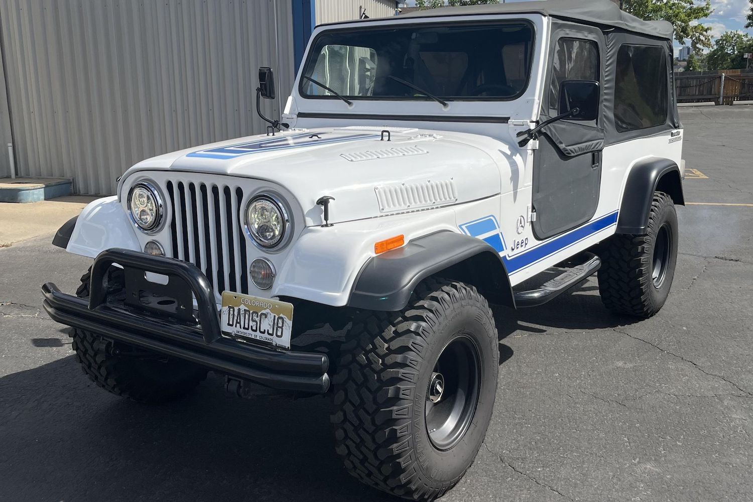 The Jeep grille of a white CJ-8 Scrambler 4x4 restomod with Mercedes turbodiesel badges, a car dealership visible in the background.