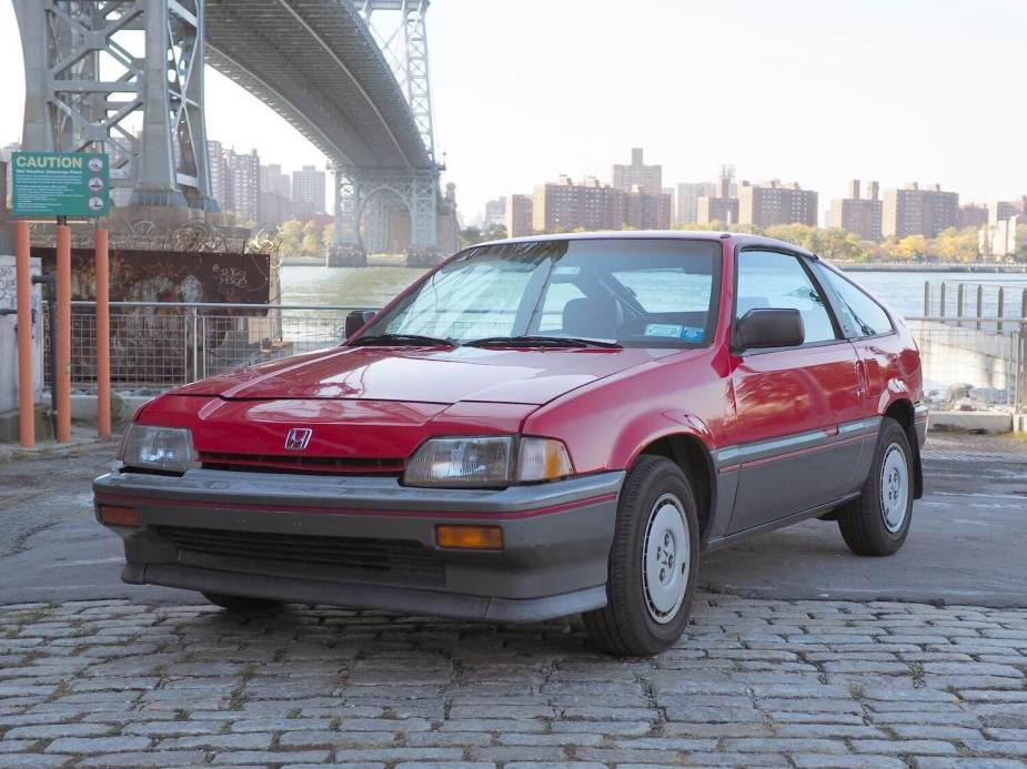 A red Honda Civic CRX coupe parked under a bridge, a city skyline visible in the background.