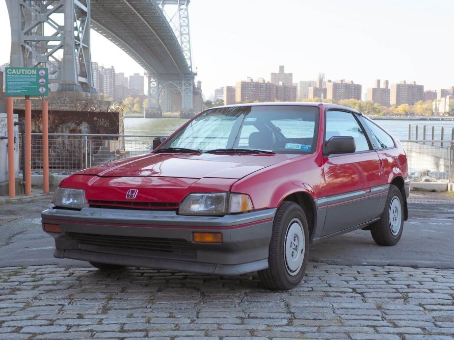 A red Honda Civic CRX coupe parked under a bridge, a city skyline visible in the background.