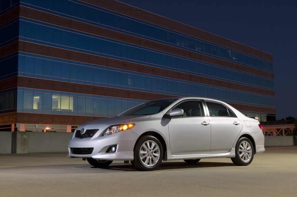 A used silver 2009 Toyota Corolla, which could have airbag and oil burning issues, poses next a high-rise building. 