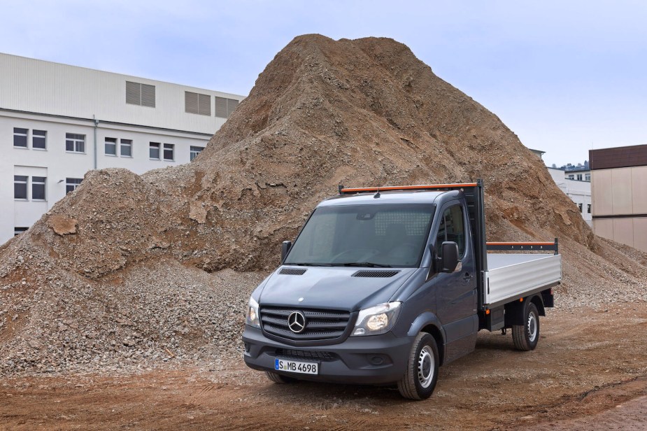 A gray Mercedes-Benz Sprinter chassis-cab with a utility bed parked in front of a sand pile.