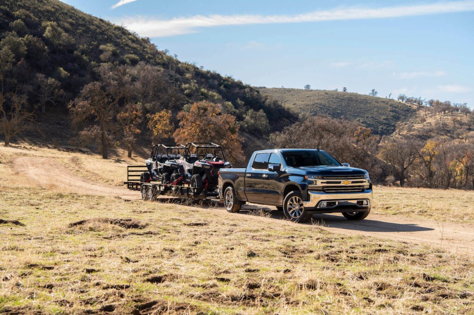Black half-ton Silverado pickup truck shows off its diesel towing capacity by pulling a trailer of four-wheelers through a field.