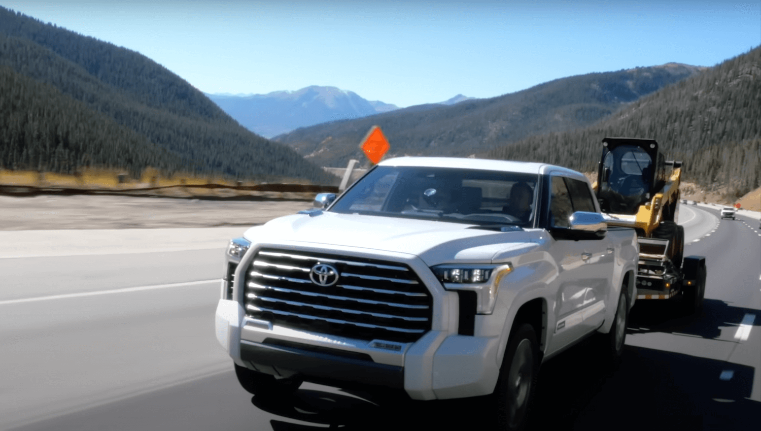 White V6 Toyota Tundra hybrid pickup truck pulls a trailer with a 10,000 skidsteer up the Rockies to test its towing mileage, mountains visible in the background.