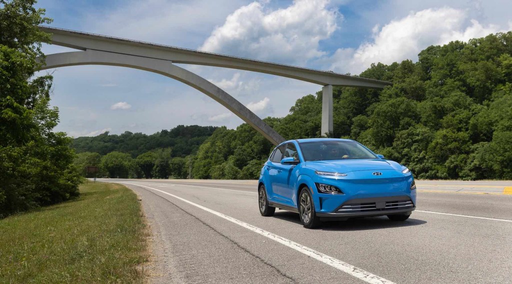 A blue Hyundai Kona Electric SUV drives past a bridge with green trees on either side of the road.
