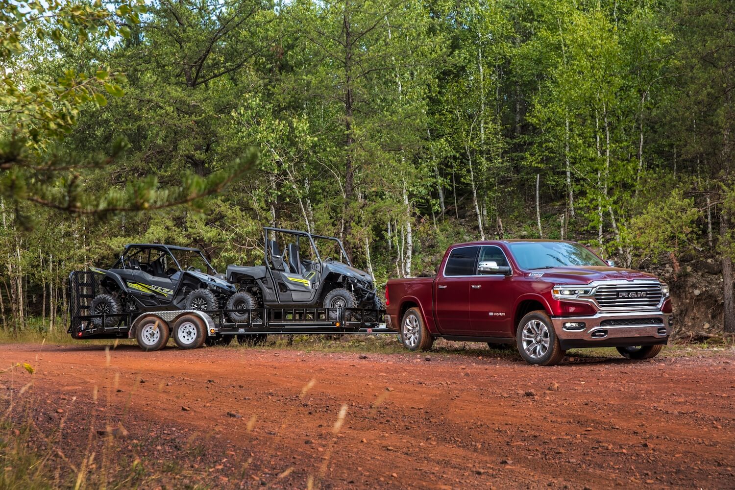 Red Ram 1500 pickup truck towing a trailer with two side-by-side 4x4s, trees visible in the background.