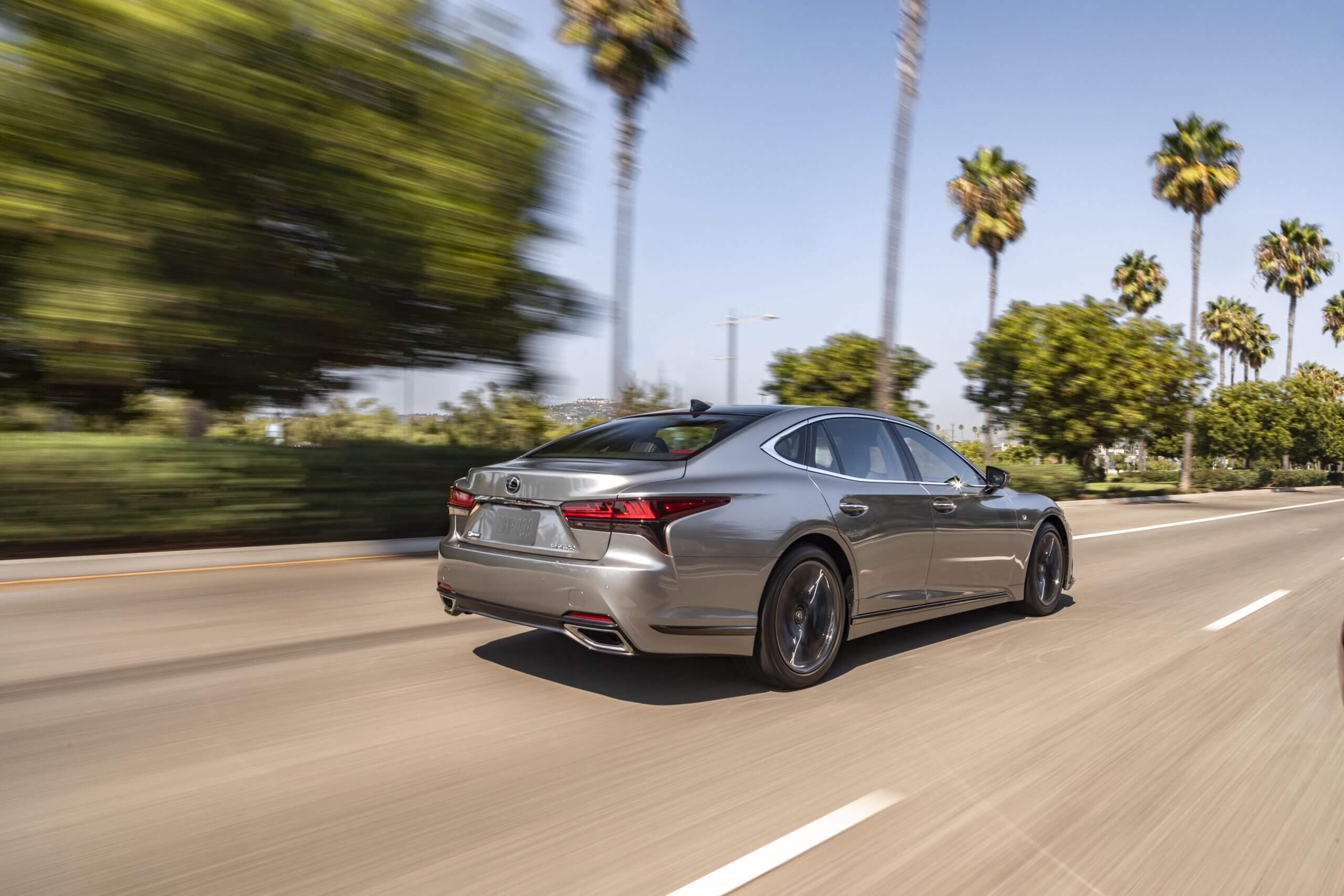 The 2023 Lexus LS speeding down a sunny road with trees.