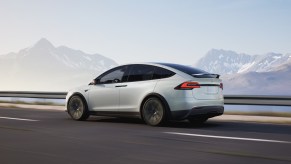Rear view of a white Tesla Model X driving down a road with mountains in the background.