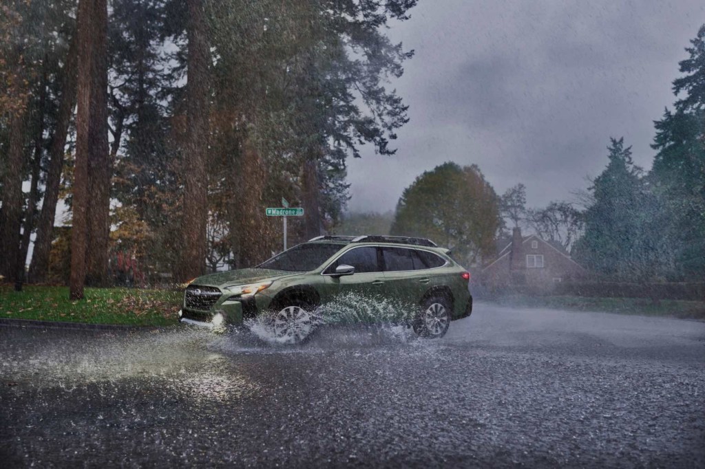 A green Subaru Outback drives through heavy rain with its headlamps illuminated. Large pine trees are in the background.