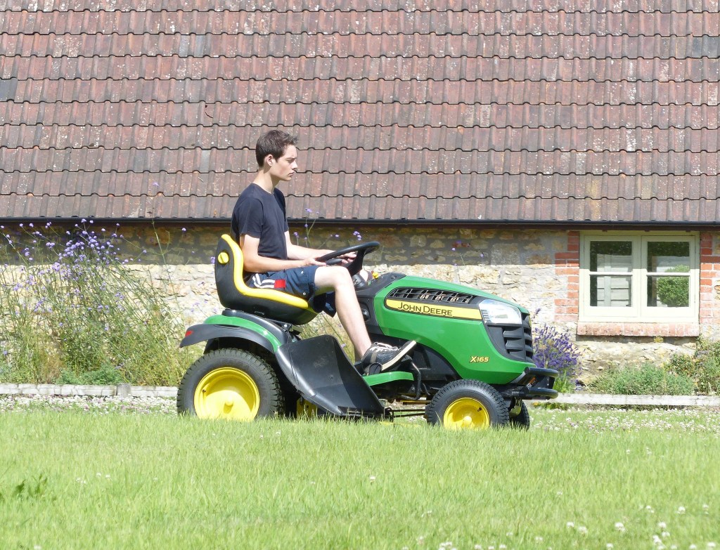 A riding lawn mower mowing grass driven by a younger person that might be in the need of removing the lawn mower blades.