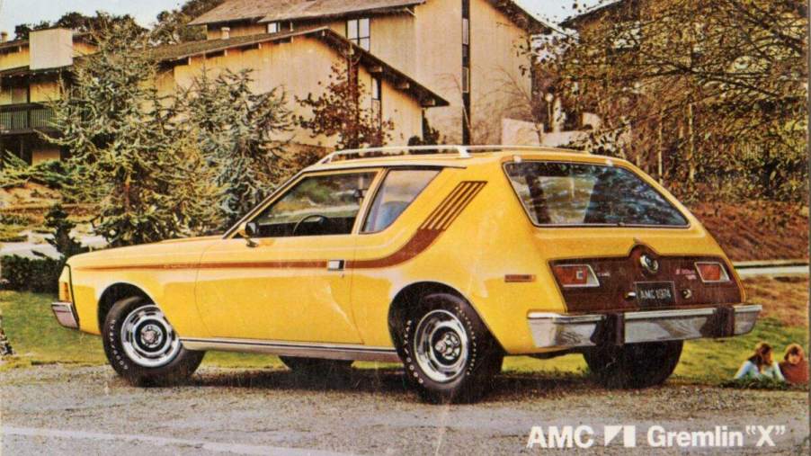 A yellow AMC Gremlin parked in front of a brown building with some tress.