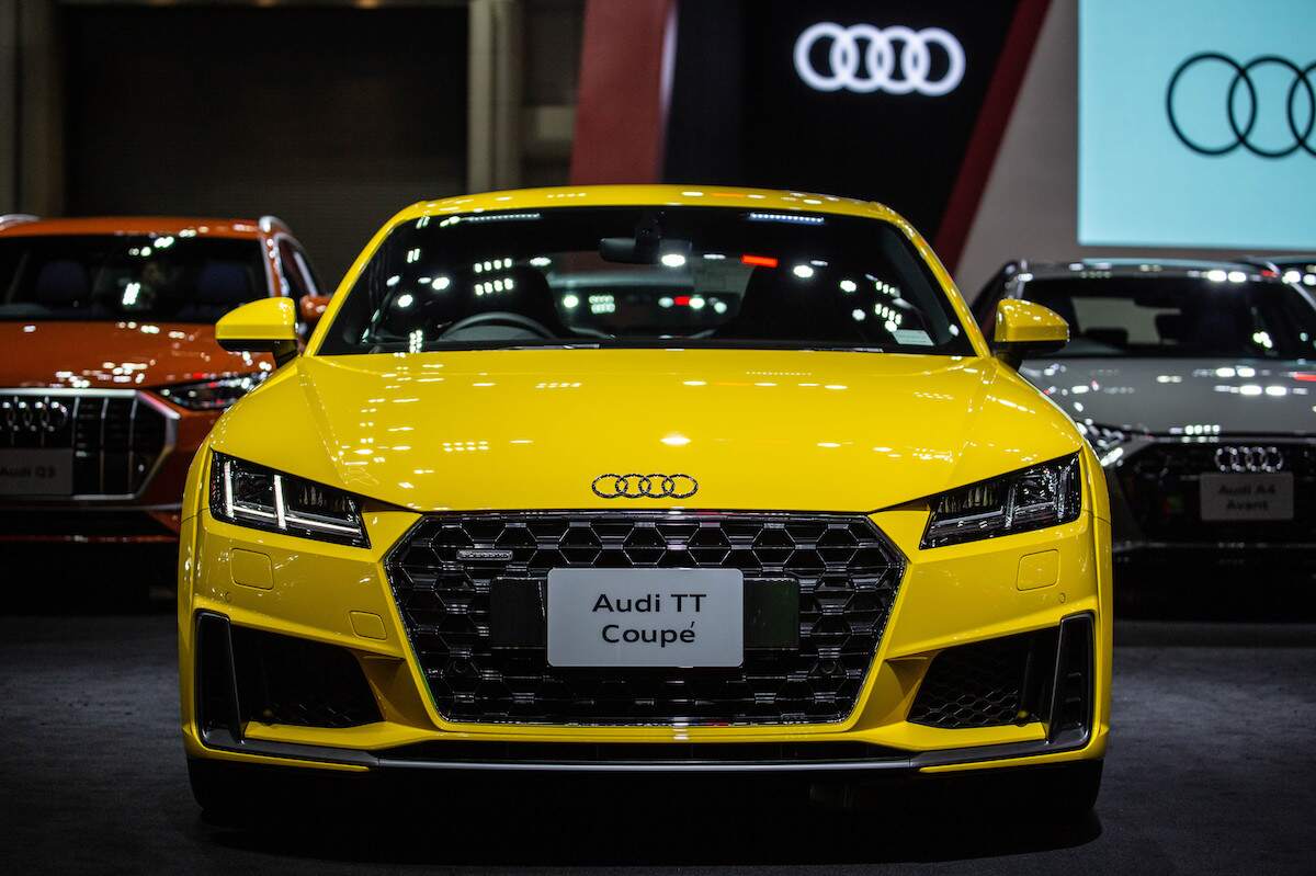 A yellow Audi TT, one of the most reliable luxury sports cars, parked indoors with the Audi logo in the back.