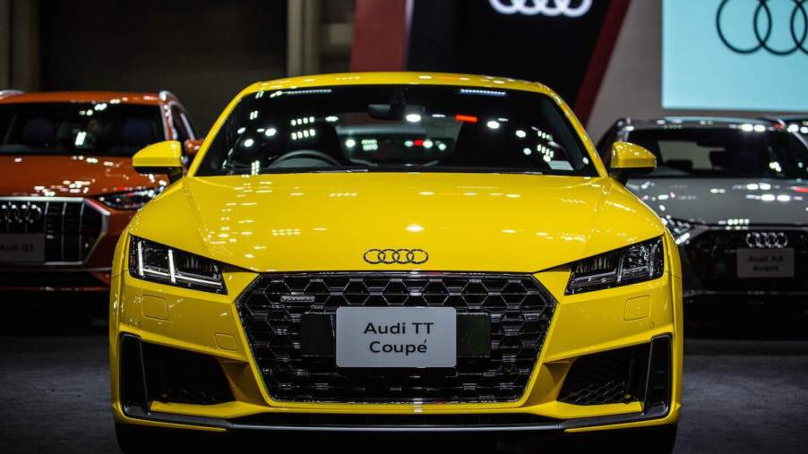 A yellow Audi TT, one of the most reliable luxury sports cars, parked indoors with the Audi logo in the back.