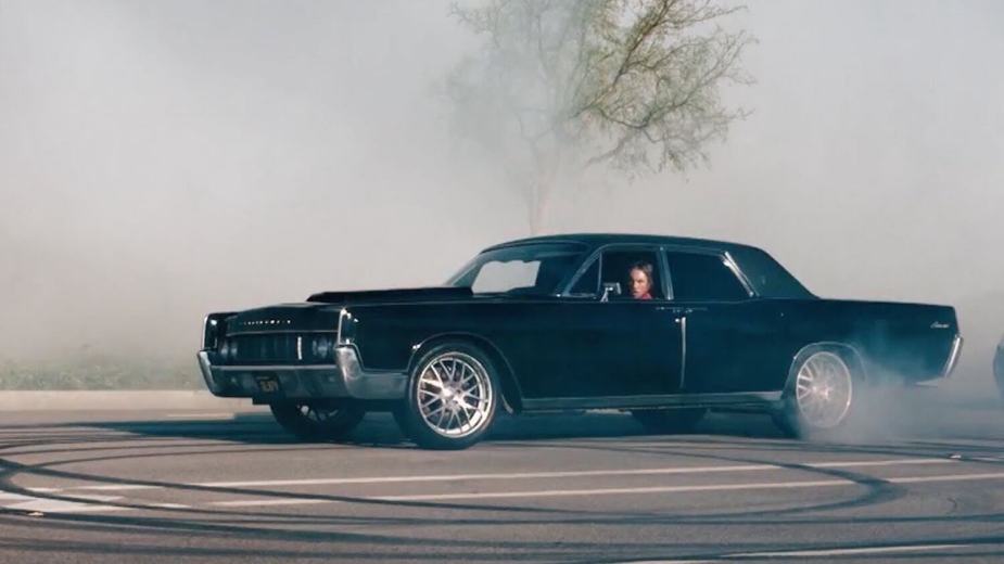 A black four-door Lincoln Continental sedan doing donuts in a parking lot surrounded by a cloud of tire smoke.
