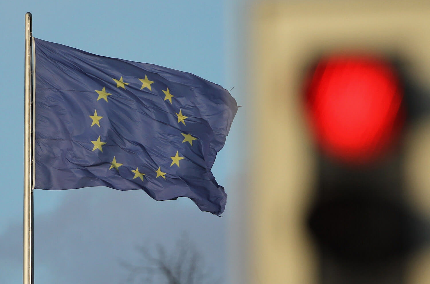 A waving blue European Union flag with a blurry traffic stoplight visible in the background.