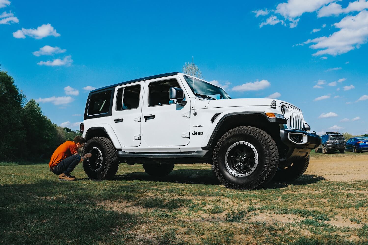 A woman crouches by a white Jeep Wrangler SUV to adjust its tire pressure, a field and blue sky visible in the background.