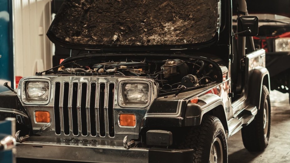 The grille of a Jeep Wrangler TJ with its hood up, parked in a garage, another SUV visible in the background.