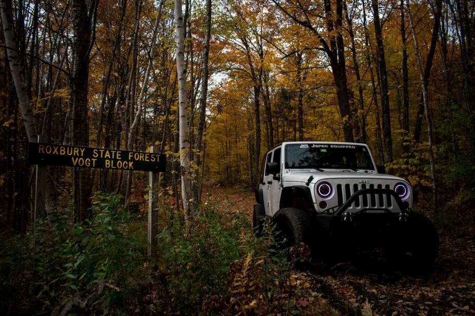 A Silver Jeep Wrangler Unlimited drives along a leaf-strewn off-road trail in the Ground Mountain State Forest in Vermont, fall foliage visible in the background.