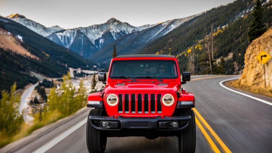 A head on view of a red Jeep Wrangler driving down a road with a mountain scenery in the background.