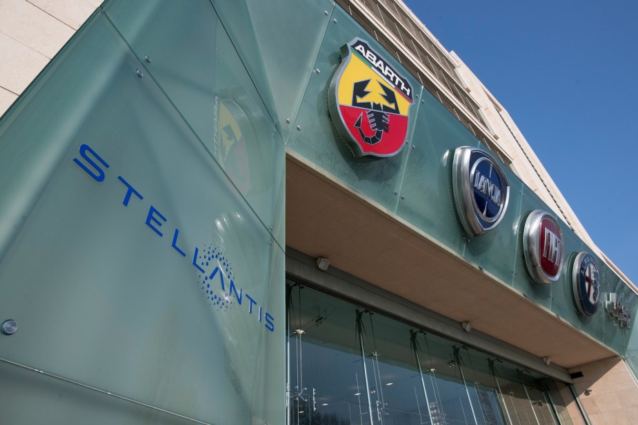 Closeup of the Stellantis logo, below the Abart, Lancia, Fiat, Alfa Romeo, and Jeep logos on an office building in Italy, the blue sky visible in the background.
