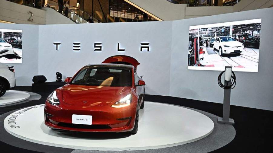 A red Tesla Model 3 on display in front of a grey wall with Tesla written on it and a screen with a video playing.