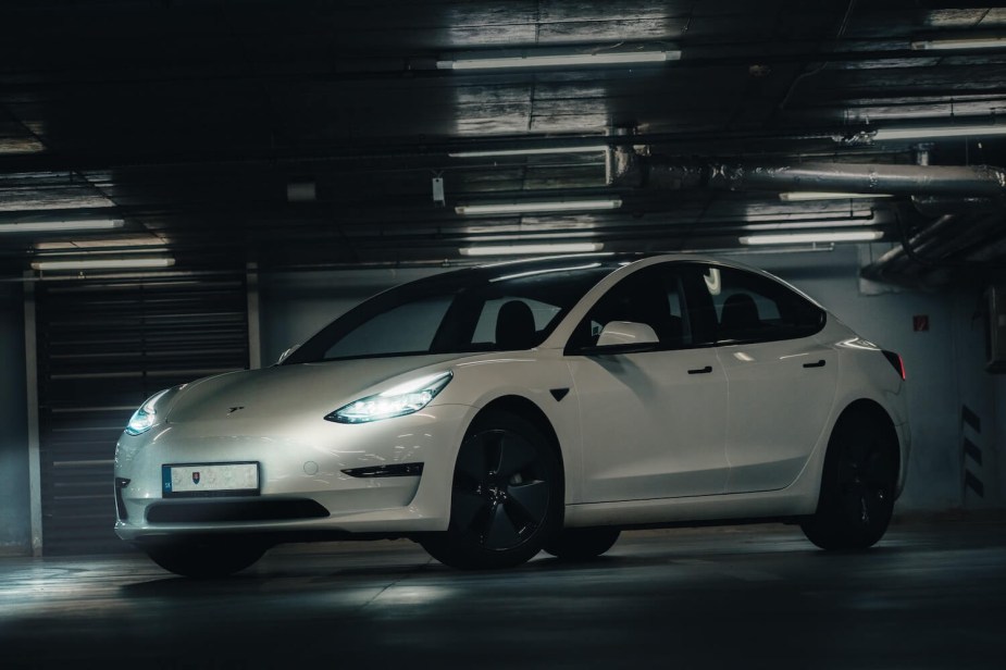 A white Tesla Model 3 is parked in an underground garage, its headlights illuminating the concrete.