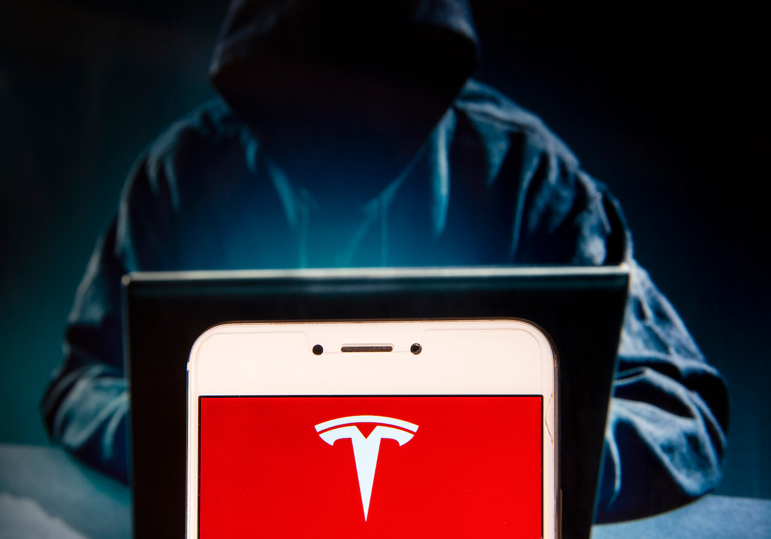 A person in a hoody types on a laptop in the background, while the Tesla logo is visible on a cellphone in the foreground.