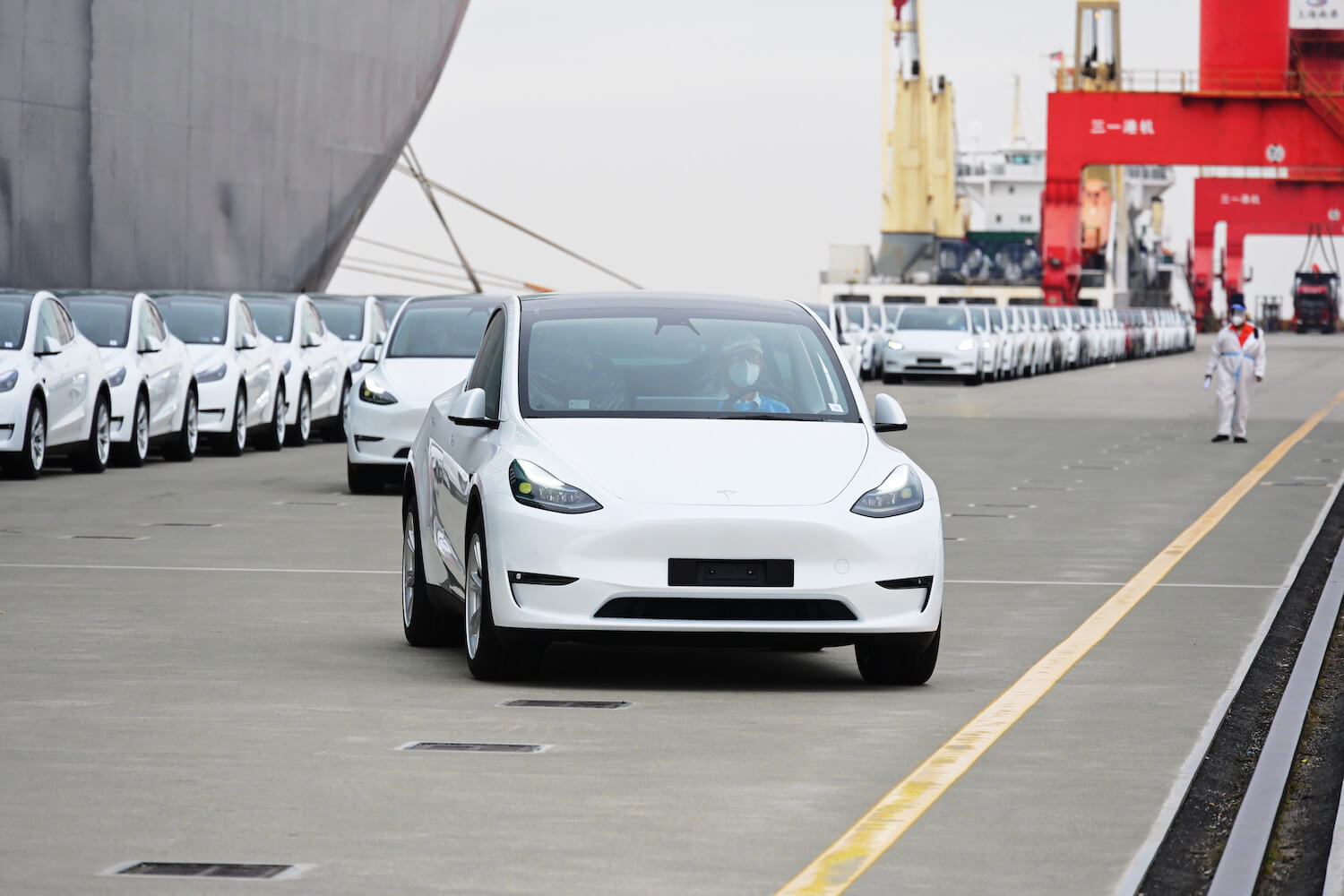 A white Tesla Model Y electric SUV in a harbor being exported to China for foreign sales, a transport ship visible in the background.
