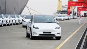A white Tesla Model Y electric SUV in a harbor being exported to China for foreign sales, a transport ship visible in the background.